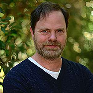 Rainn Wilson standing with his arms crossed in front of greenery